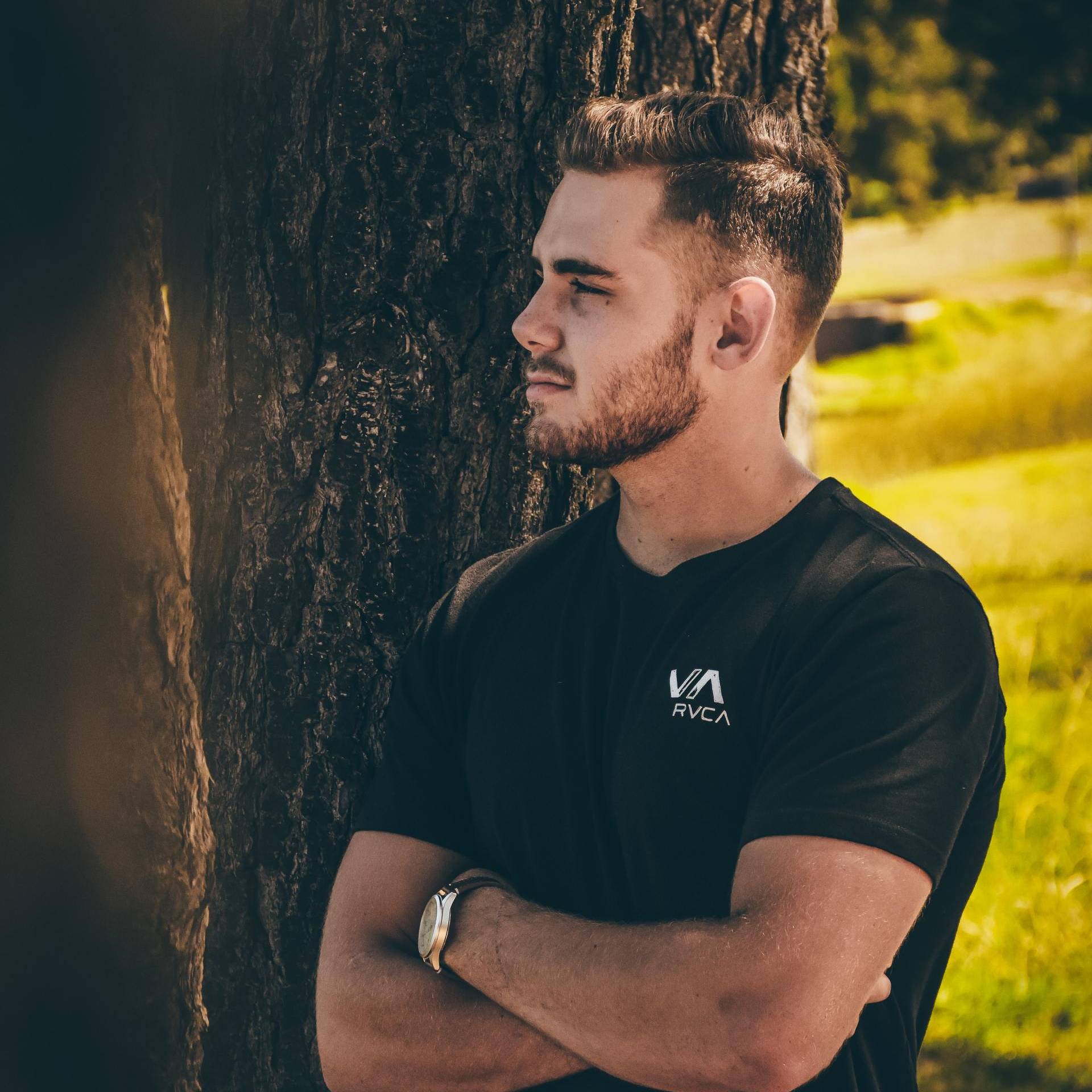 Man in his early twenties standing next to a tree wearing a black t-shirt looking out the left of the frame. 