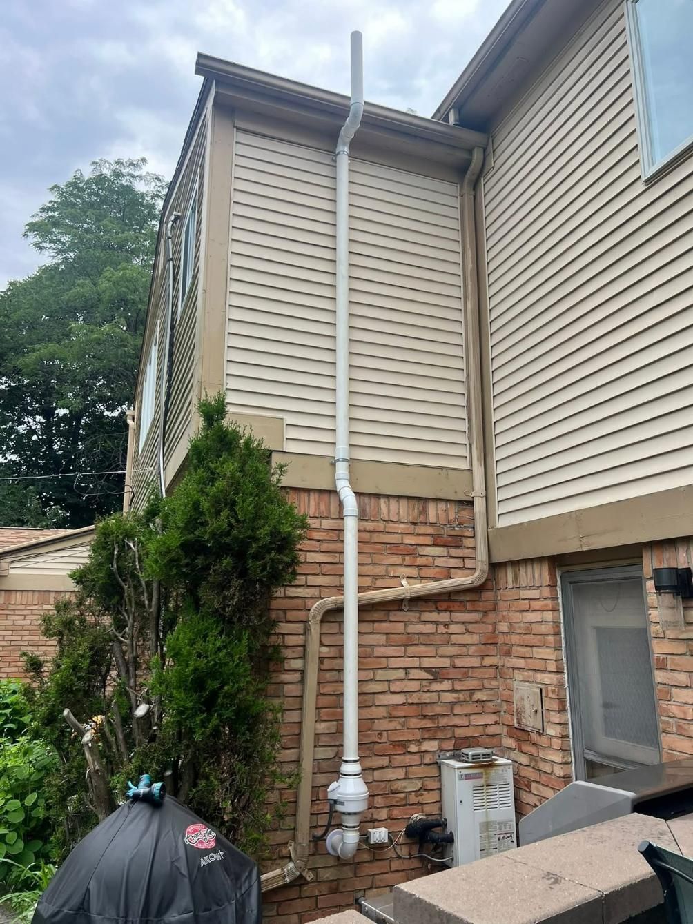 White pipe runs vertically down a brick and beige-sided building exterior. Trees and the sky are visible.