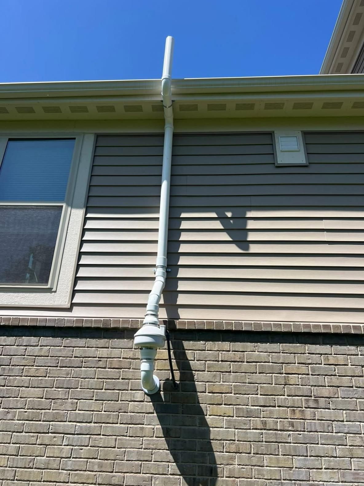 White downspout attached to a gutter on a house with tan siding and a brick foundation.
