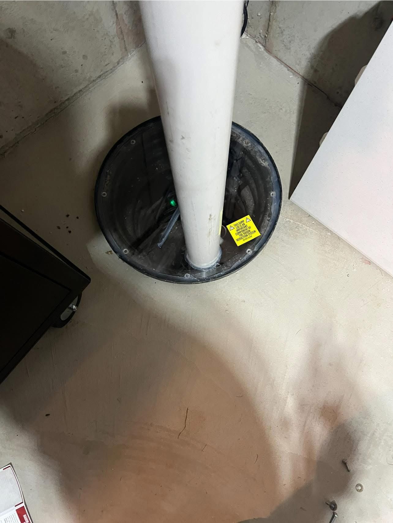 A sump pump in a concrete basement corner. A white pipe rises from a black, circular basin on the floor.
