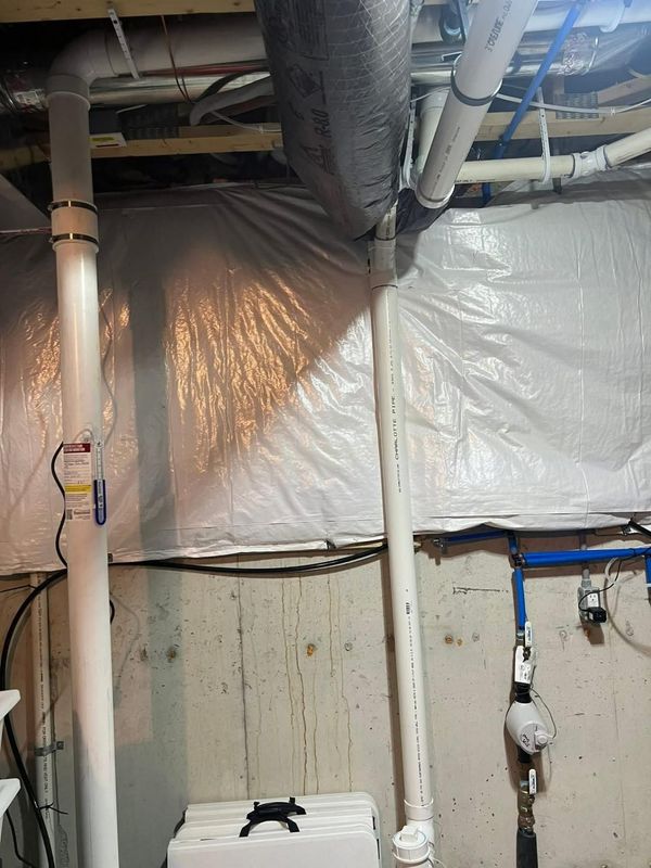 Pipes and a flexible duct against a wall. A white wrap covers the wall. Plumbing is visible.