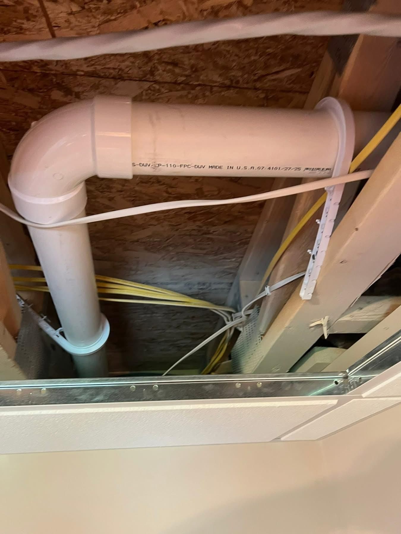 White PVC pipe system in a ceiling cavity with electrical wires and wooden framing.