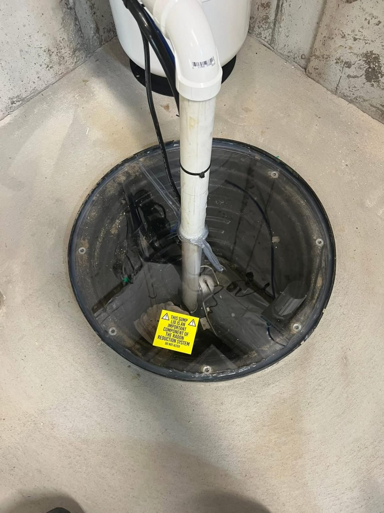 A sump pump in a round black pit in a basement corner, with a white pipe leading up.