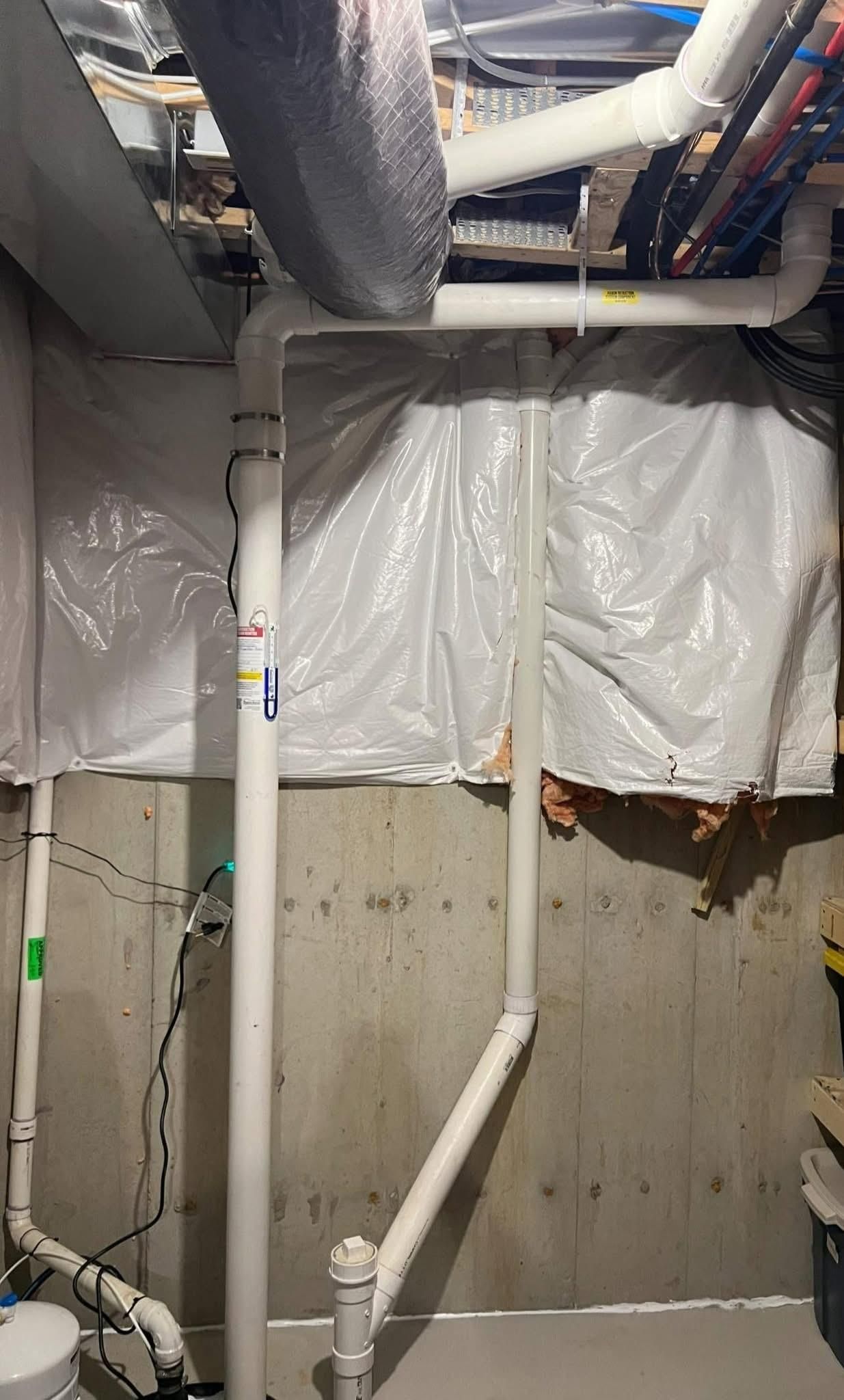 White pipes in a basement, running along a concrete wall, covered in plastic sheeting.