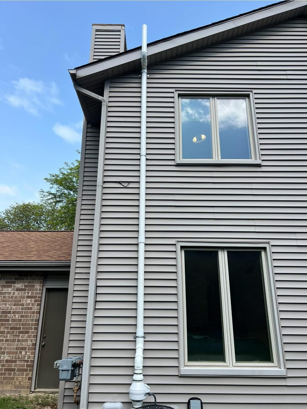Tall white pipe mounted on gray house siding. Windows visible, blue sky backdrop.