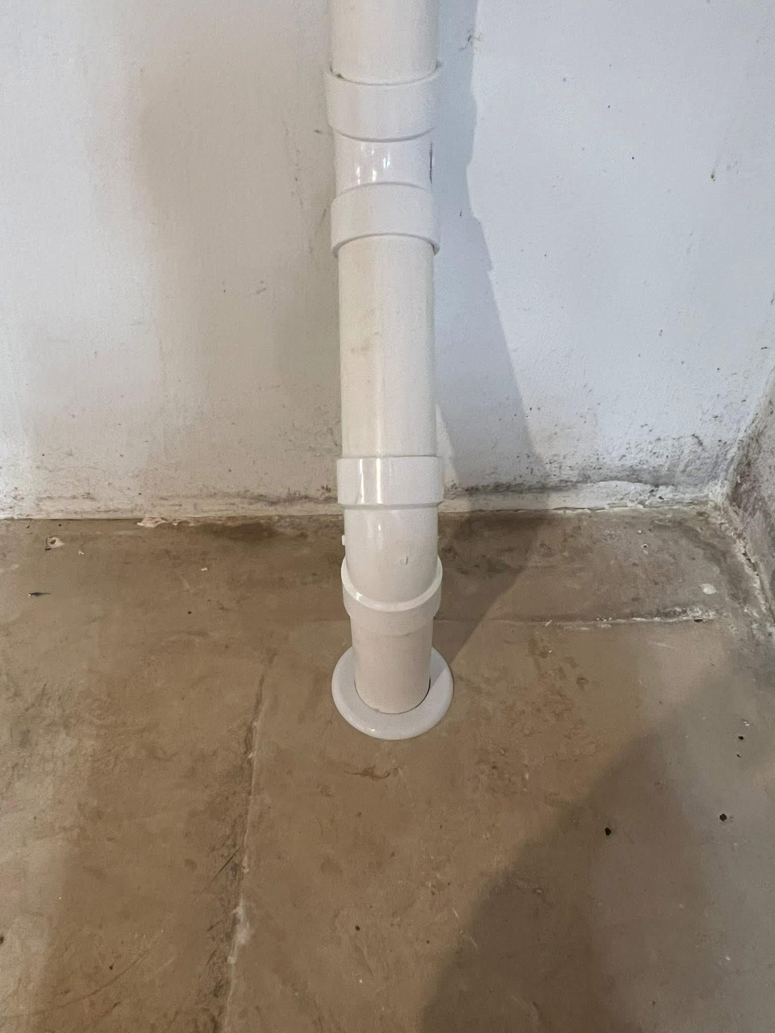 A vertical white drain pipe on a concrete floor against a white and gray wall.