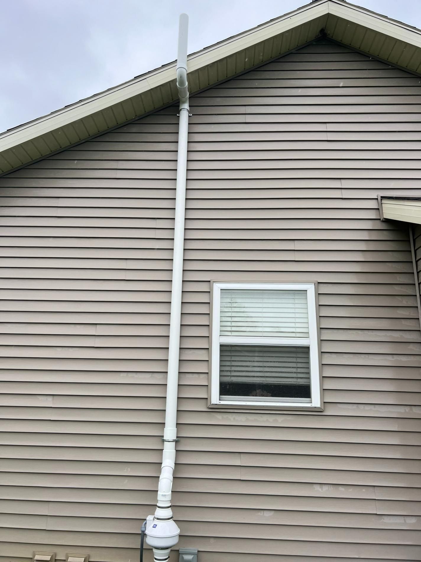 A tall white antenna is mounted to the side of a tan house, near a window.