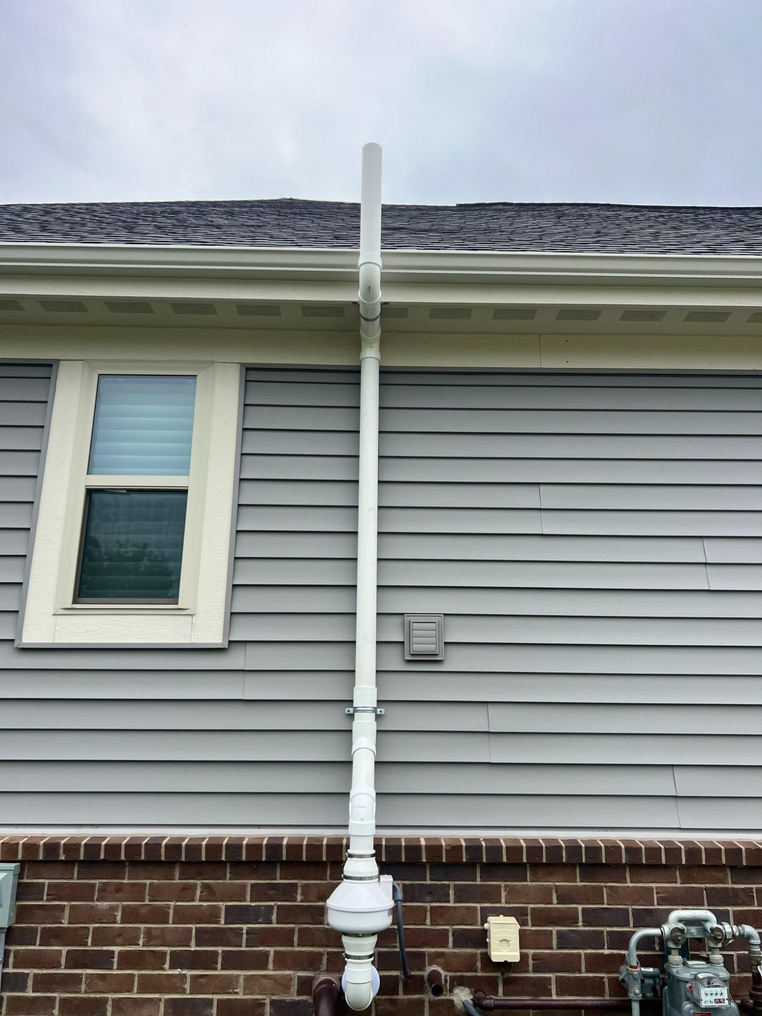 White pipe mounted on a gray house, with a gas meter and window visible. Cloudy sky.