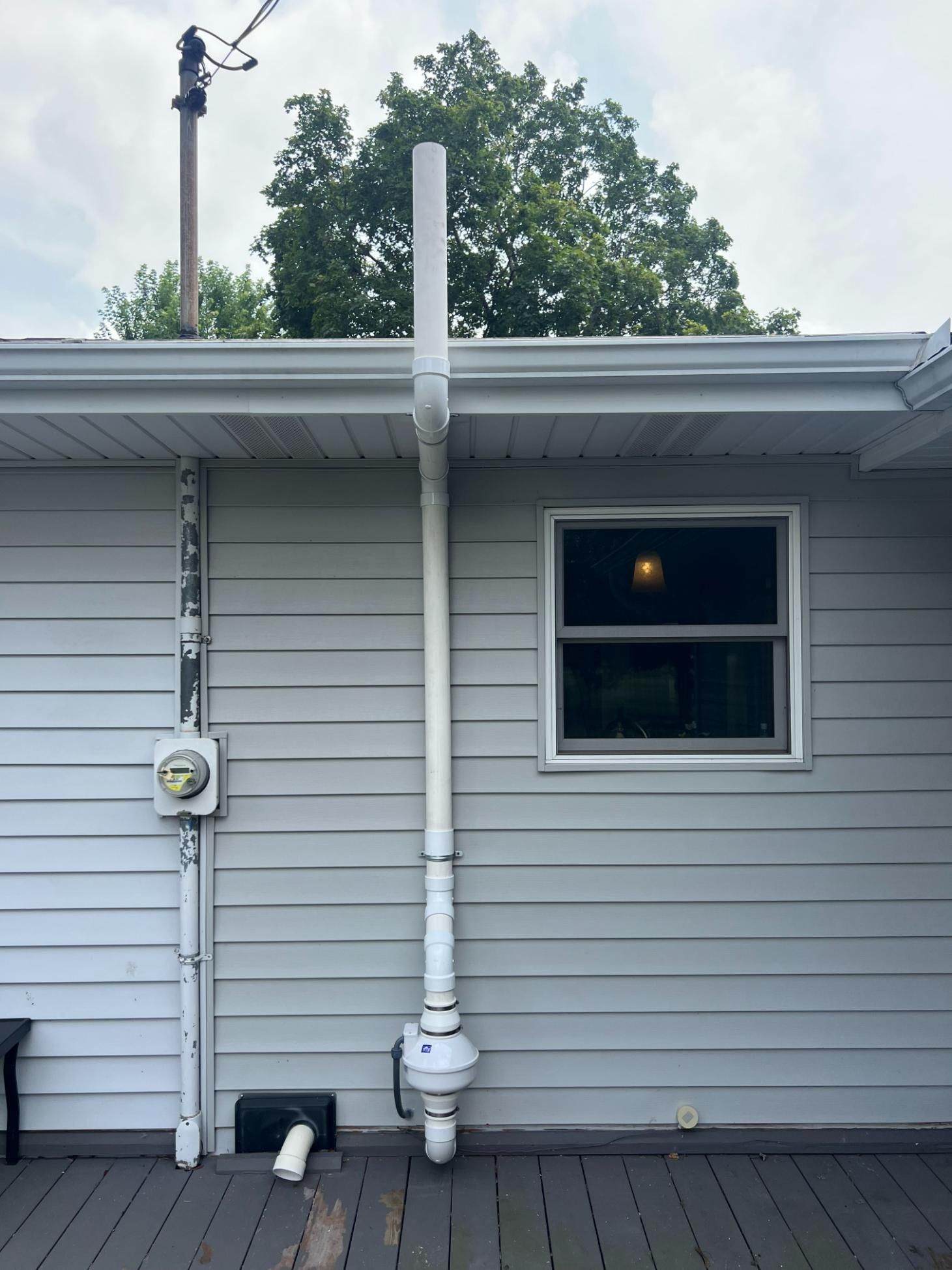 A long white pipe vents from a house's side, alongside a window, with a deck below.