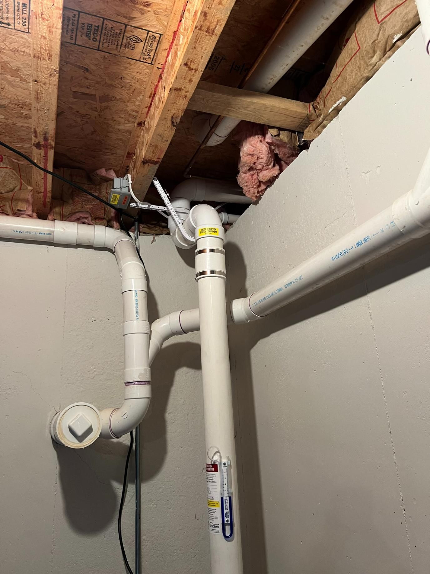 White PVC pipes in a basement ceiling against wooden beams and a gray wall.