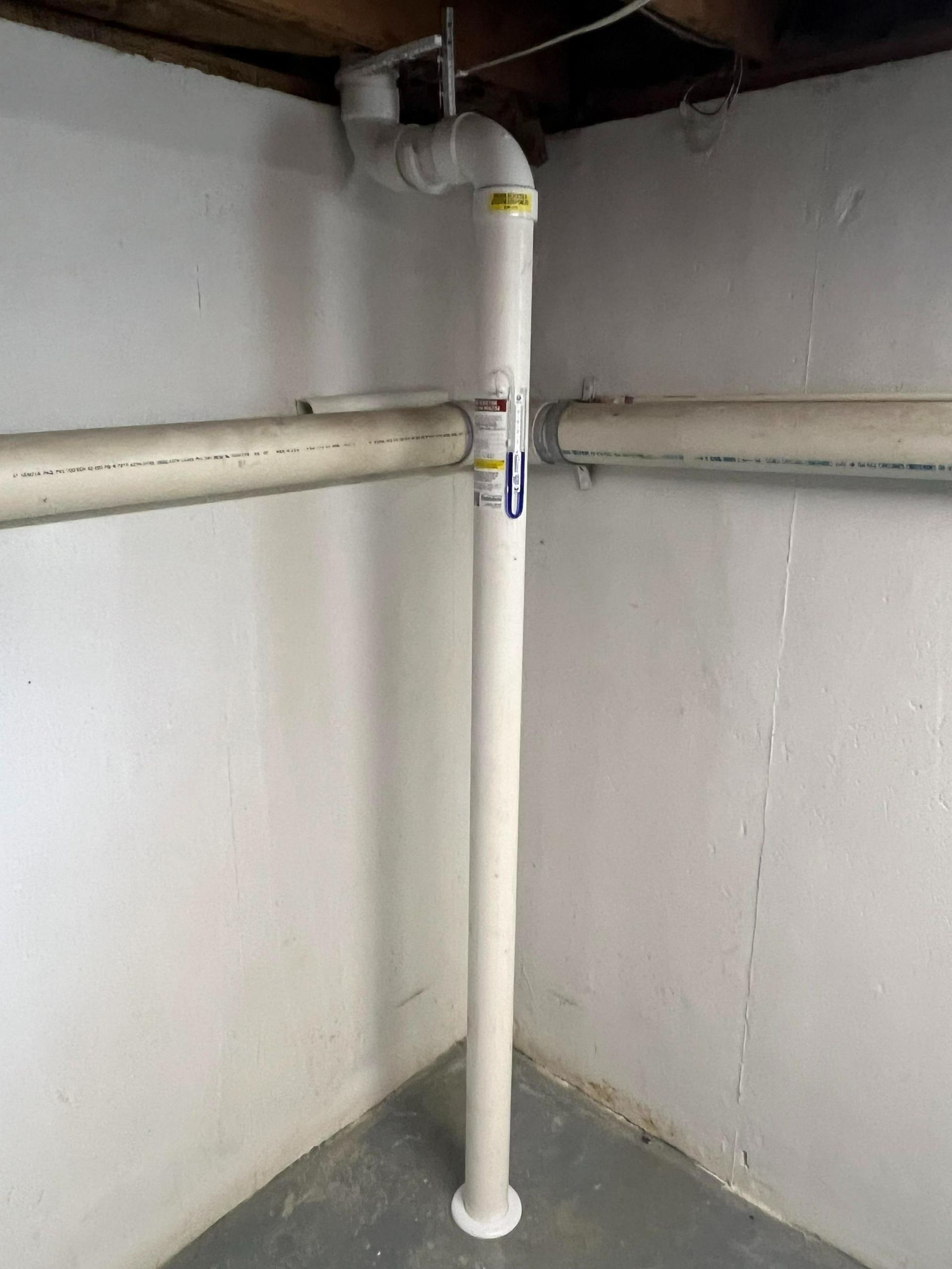 White PVC drain pipe in a basement corner; two horizontal pipes connect to it.