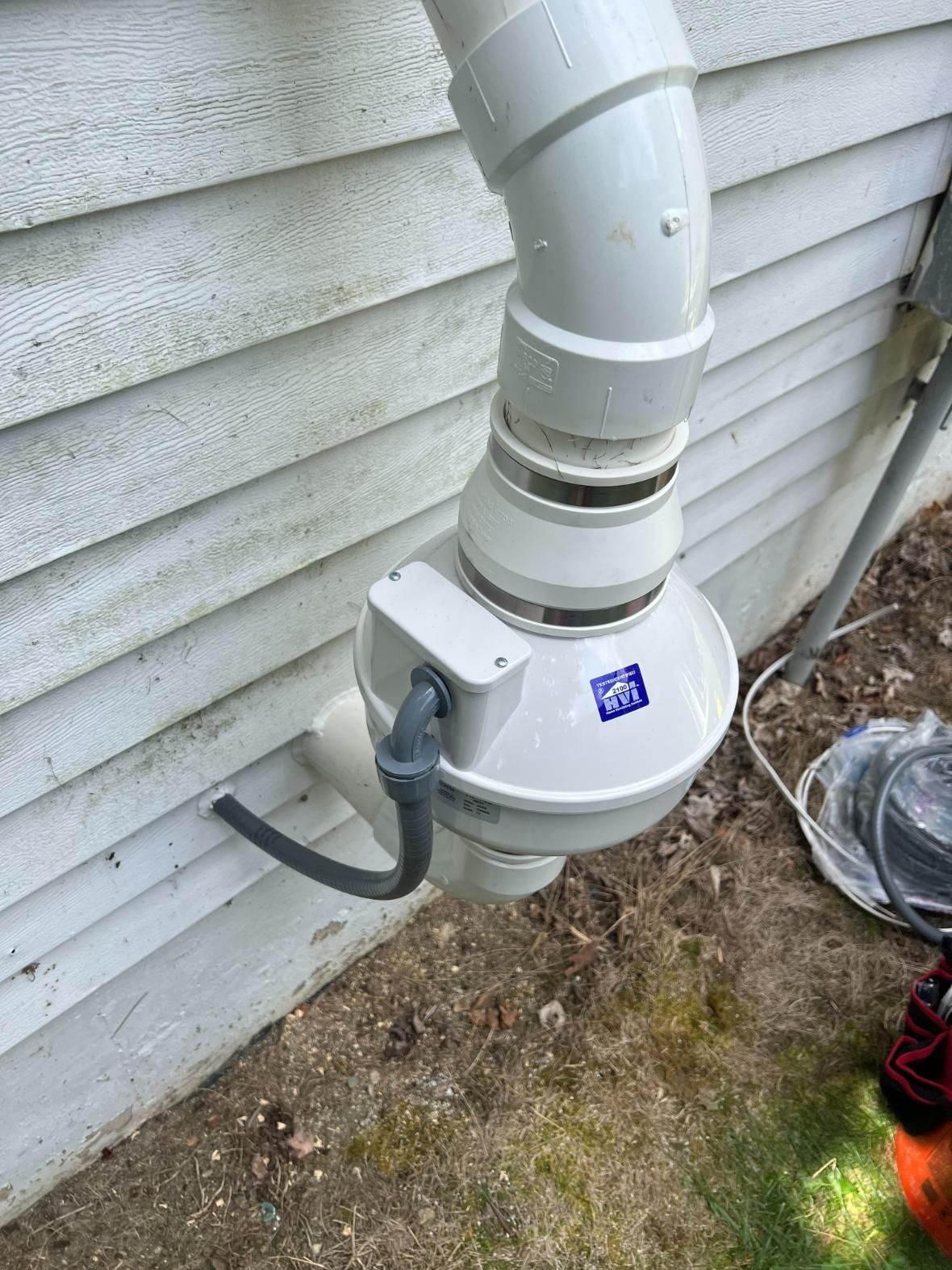White PVC pipe system on the side of a house, possibly for drainage or ventilation. Includes a small electrical component.