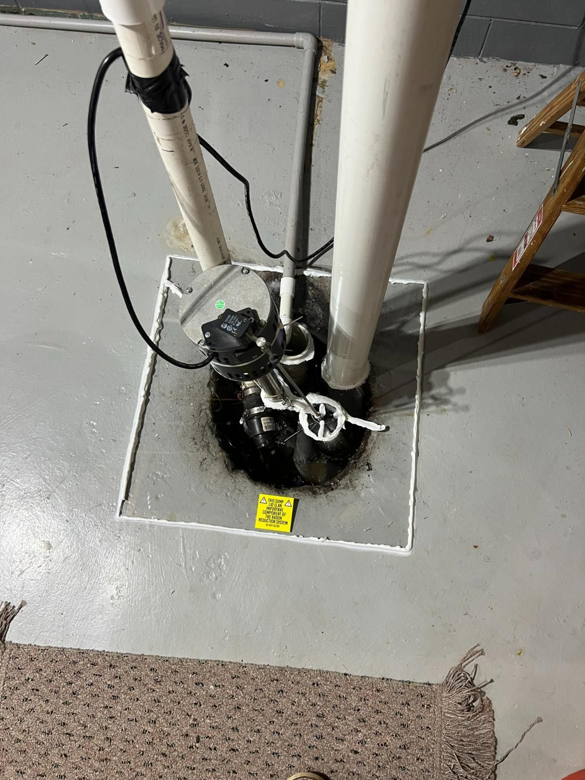 Sump pump system in a basement. Black pump, white pipes, gray floor.