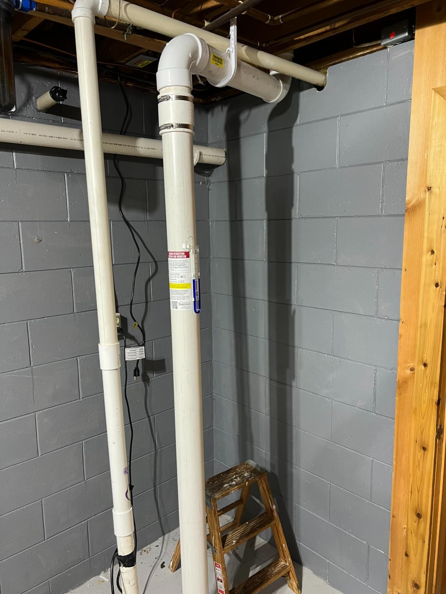 White PVC pipes against a gray cinder block wall in a basement. A small wooden ladder is at the base.
