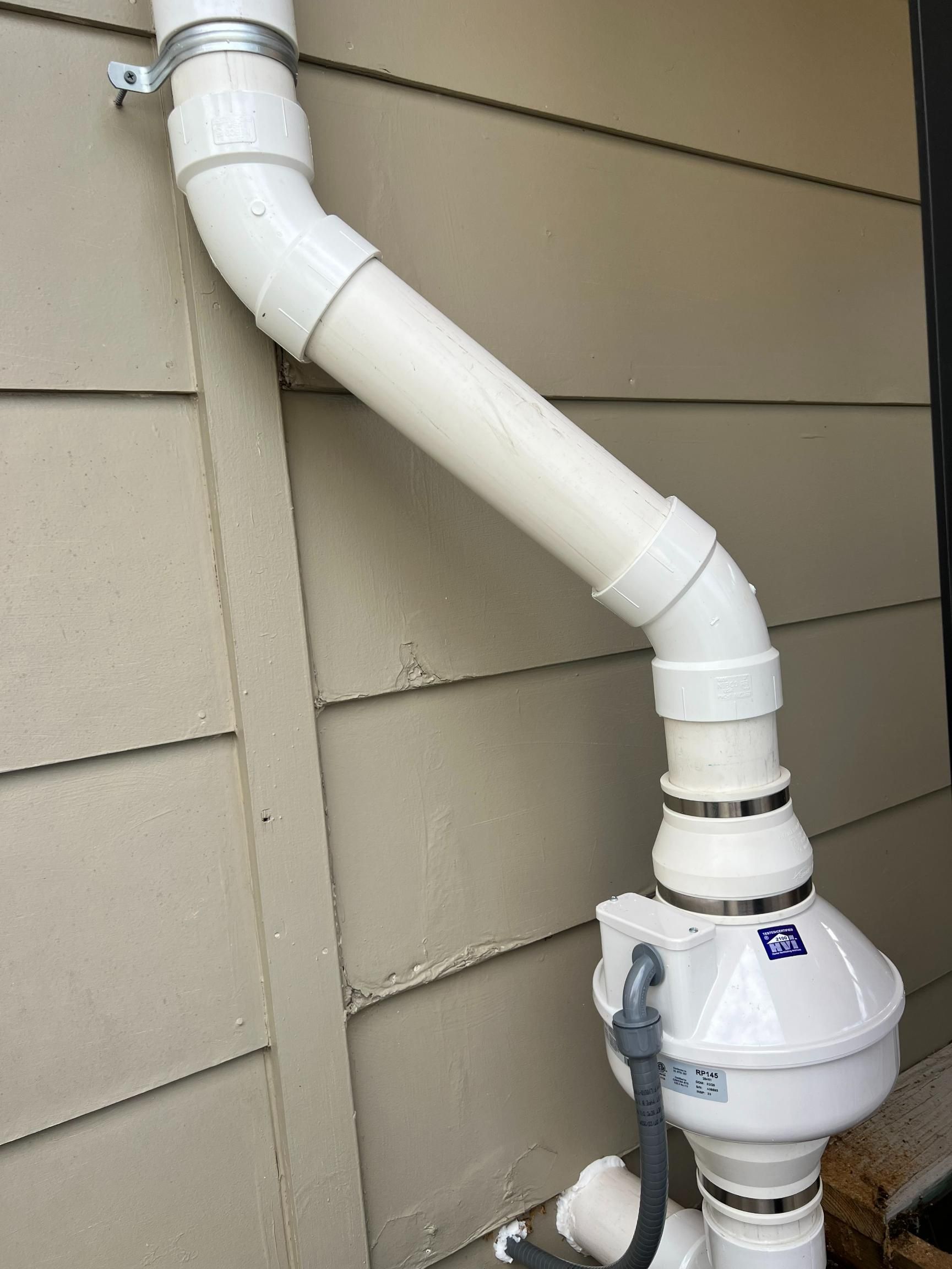 White PVC pipes attached to a beige building wall; a radon mitigation system with a fan is installed.