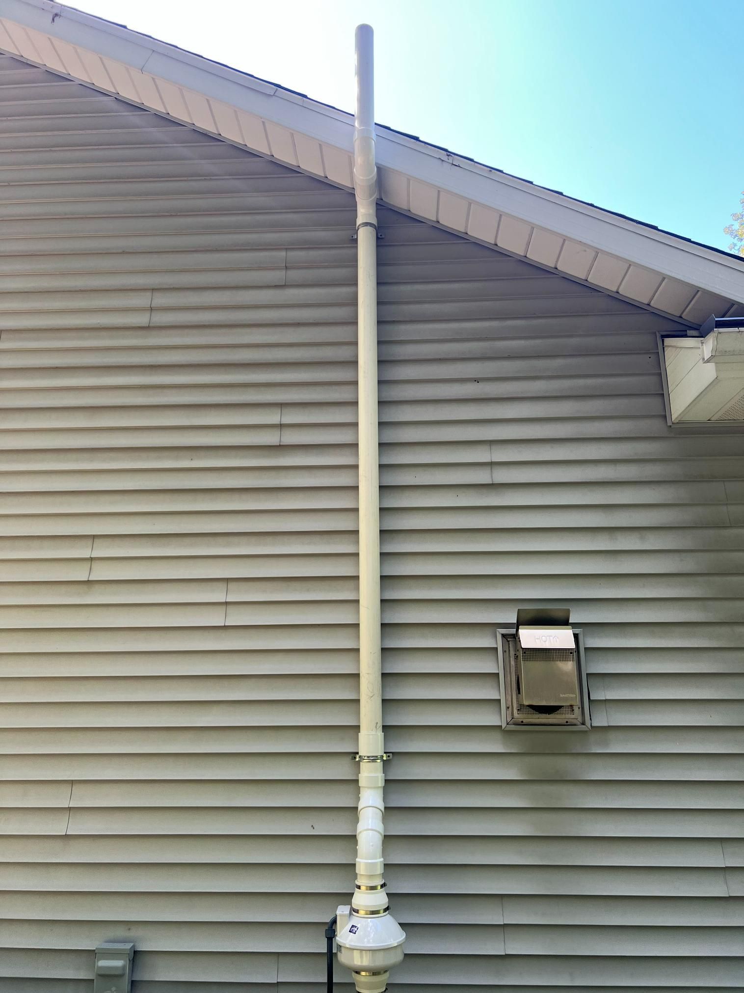 A tall, white pipe runs vertically up a light gray house siding. A vent is visible on the right.