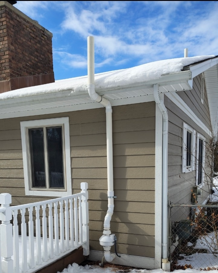 Pipes and a flexible duct against a wall. A white wrap covers the wall. Plumbing is visible.