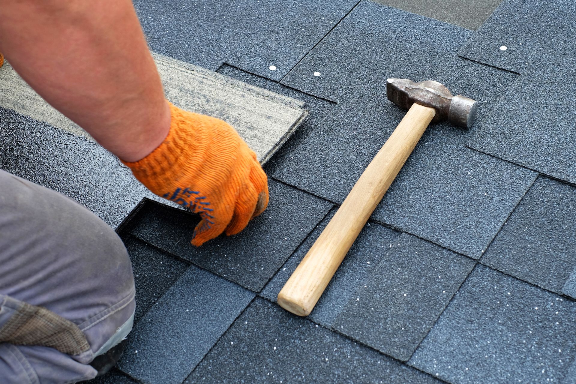 A worker in an orange glove installs dark gray asphalt roof shingles next to a hammer on a sloped roof.