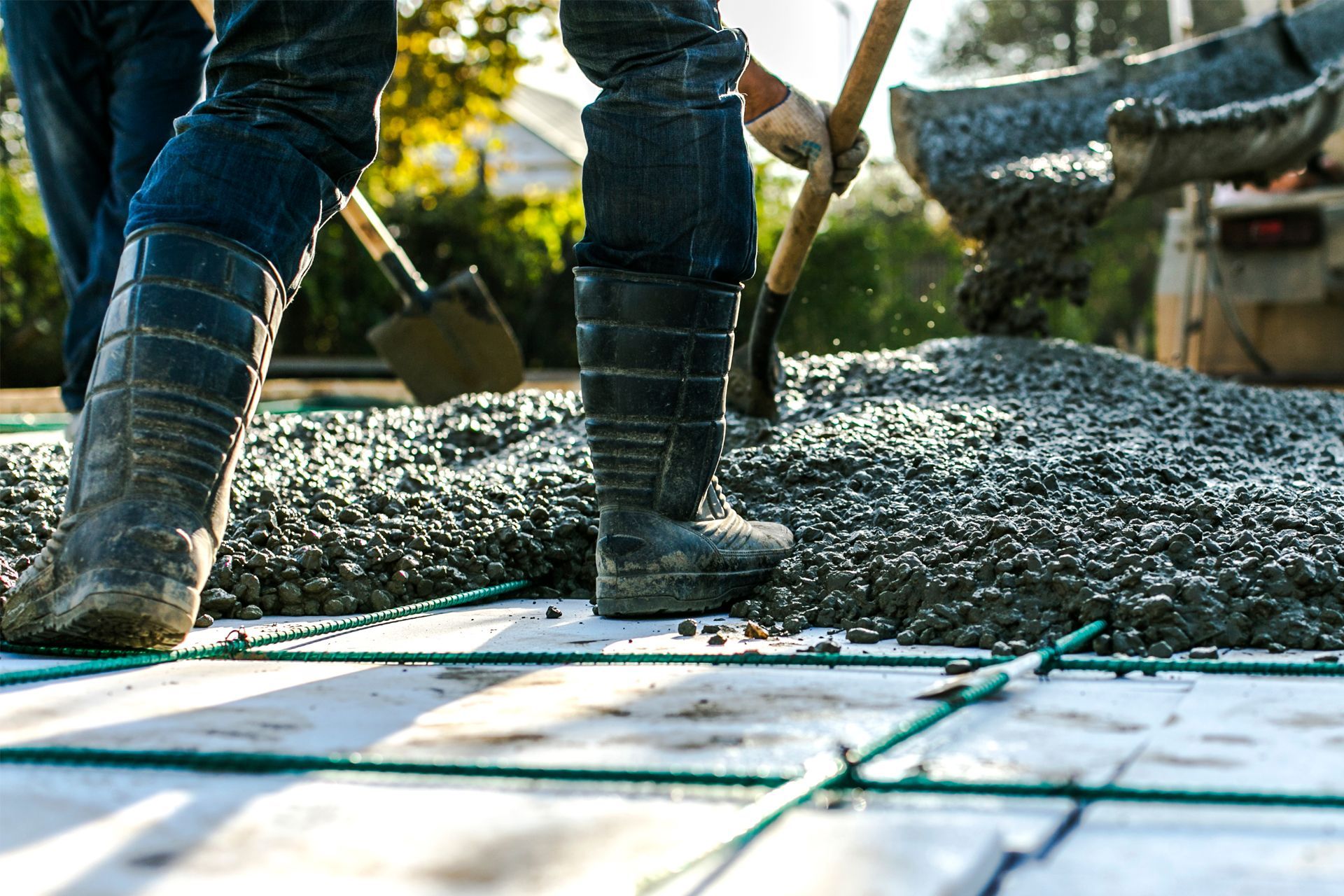 Construction workers wearing rubber boots use shovels to spread wet concrete onto a grid of wire mesh.