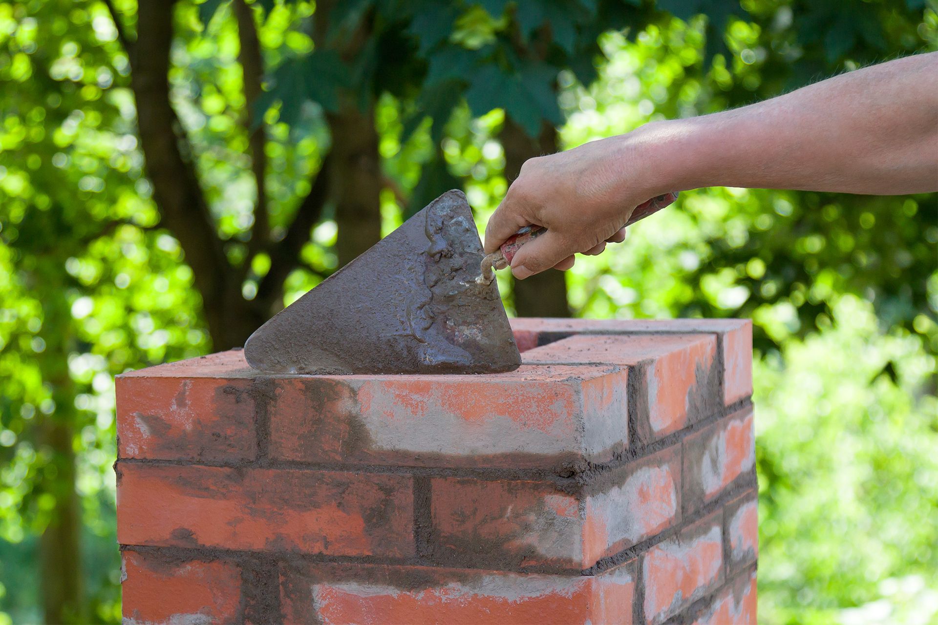 A hand uses a metal trowel to apply wet mortar onto a red brick chimney structure, with green trees in the background.