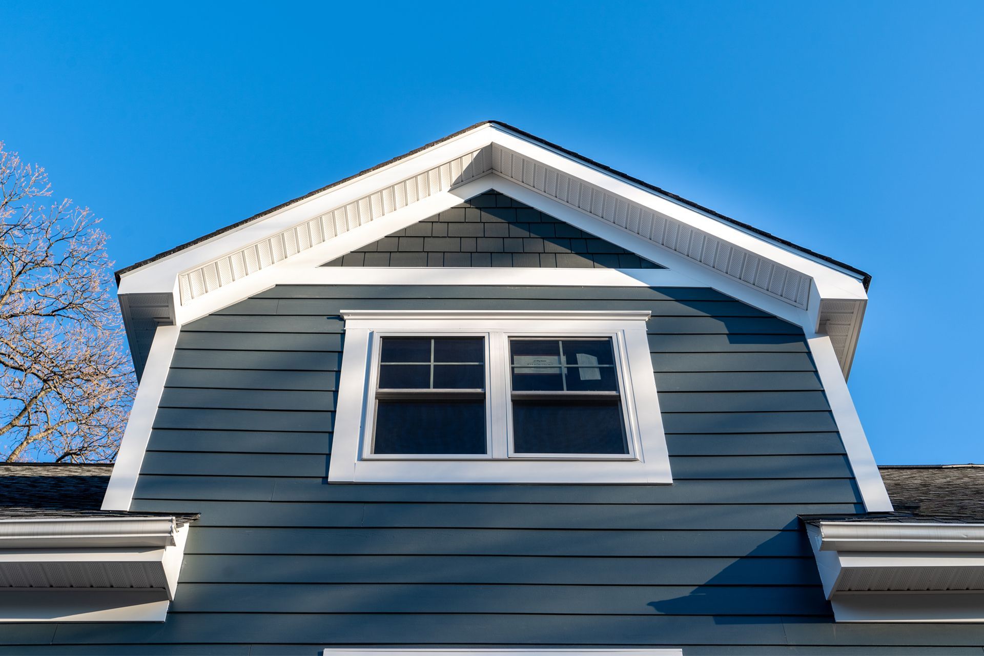 A close-up view of a blue-gray gabled dormer with a double-hung window against a clear blue sky.