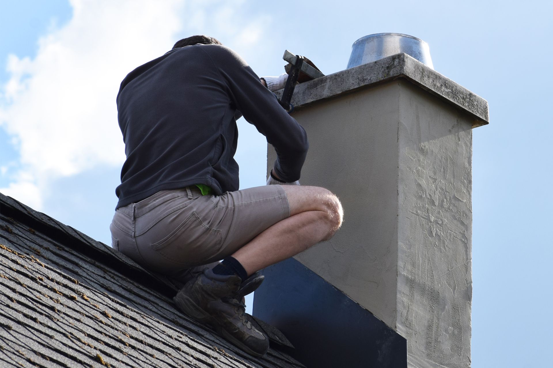 A person wearing a dark shirt and shorts squats on a sloped shingled roof, performing maintenance on a concrete chimney.