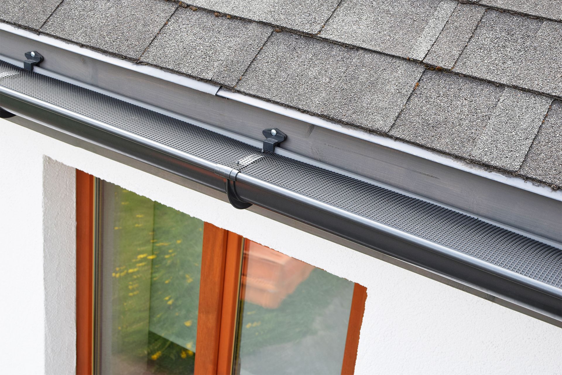 Black gutter guard mesh installed over a residential roof gutter below grey shingles and above a window.