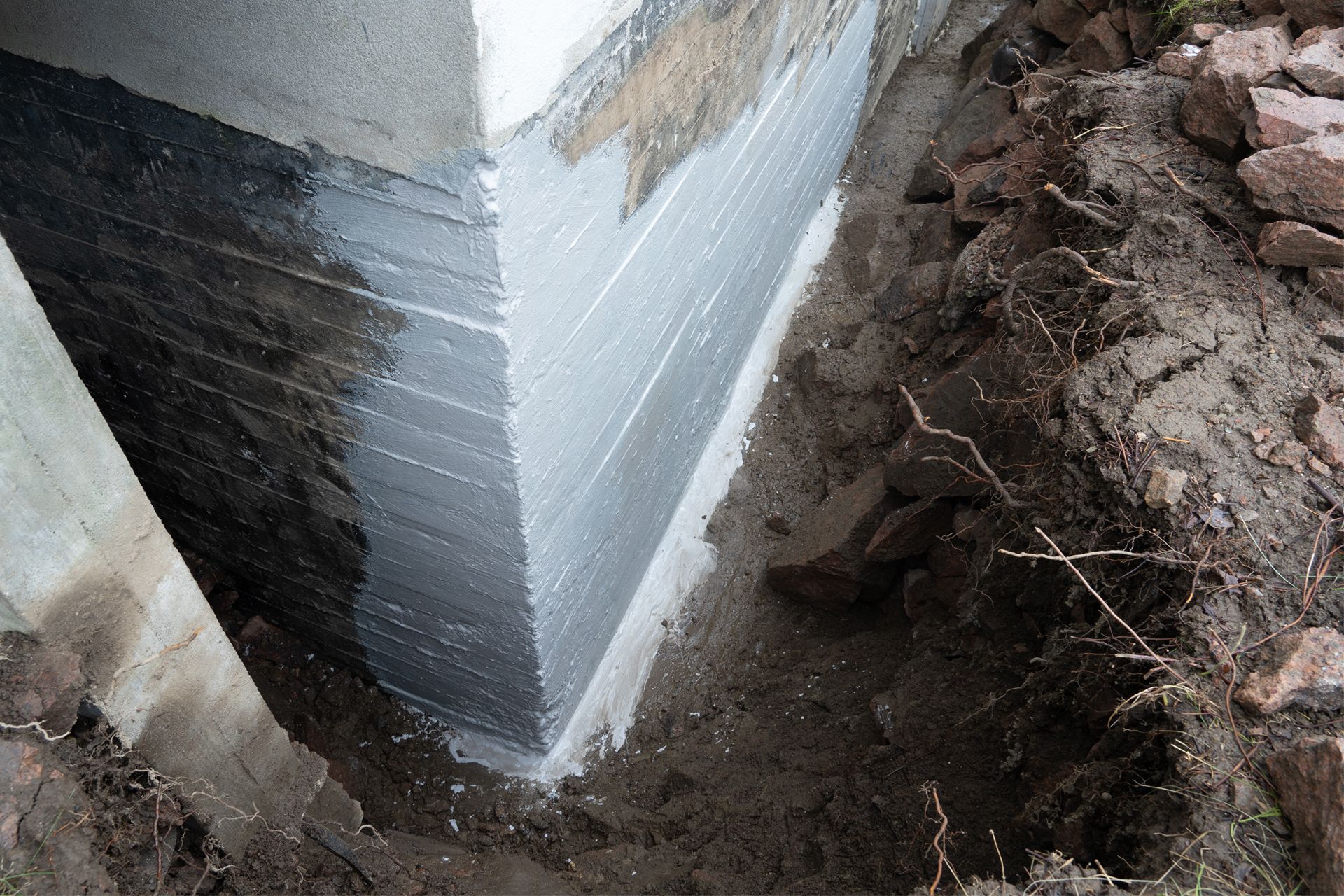 An outdoor trench dug along a concrete foundation wall, which is partially coated with grey waterproofing sealant.
