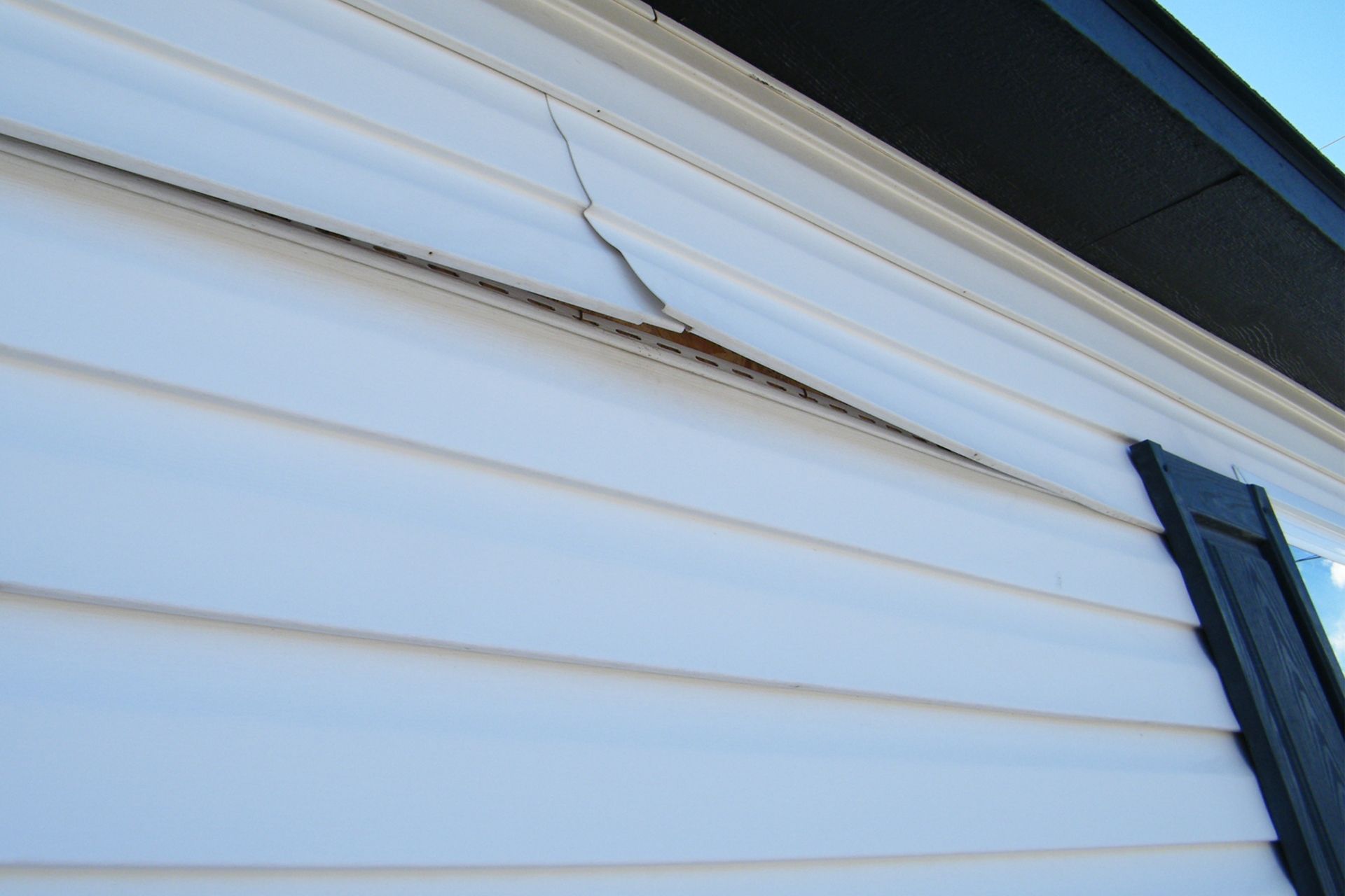 Close-up of white vinyl house siding with a vertical crack and a section of material pulling away near the eaves.