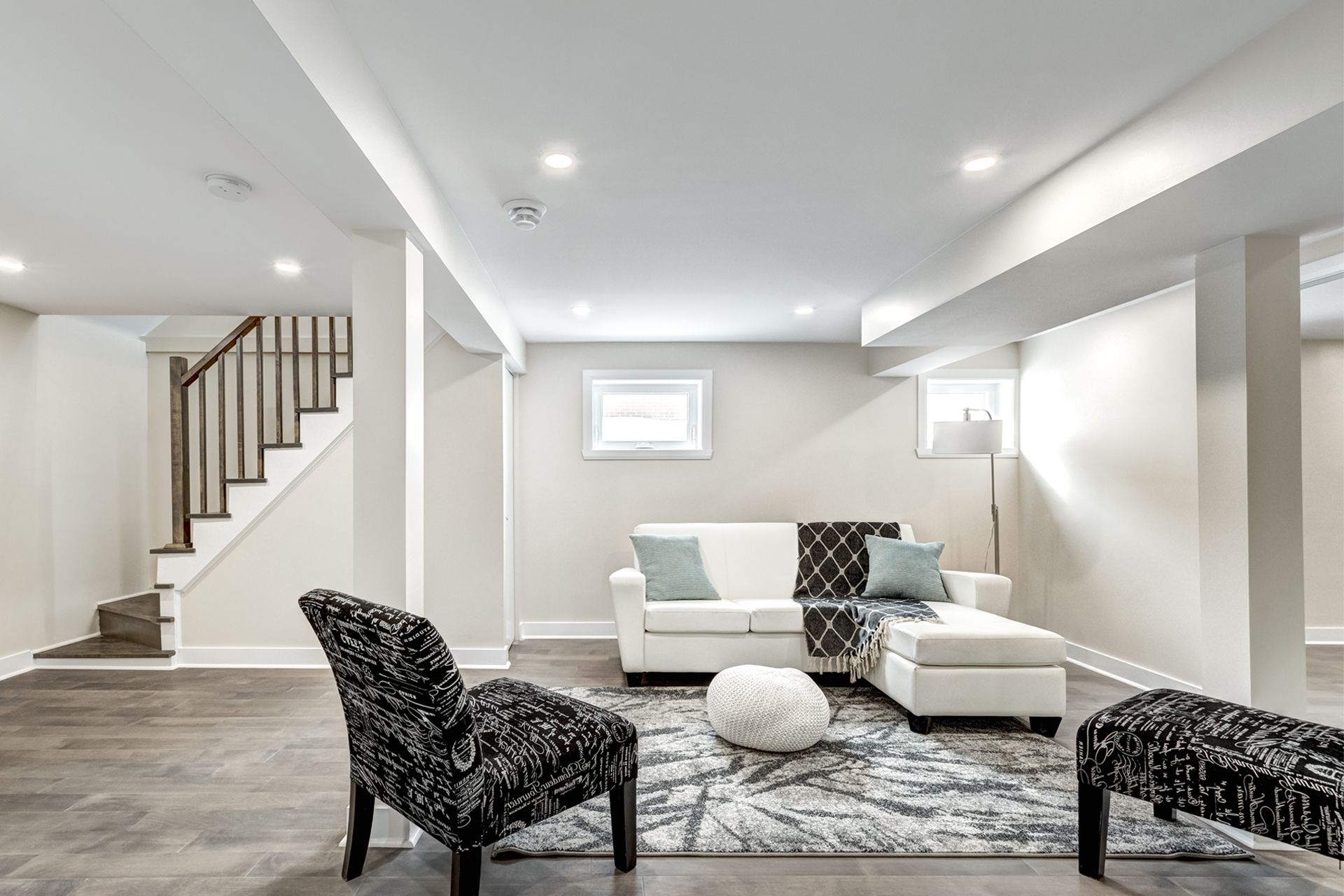 A modern, finished basement featuring a white sectional sofa, patterned armchairs, and stairs with wooden railings.
