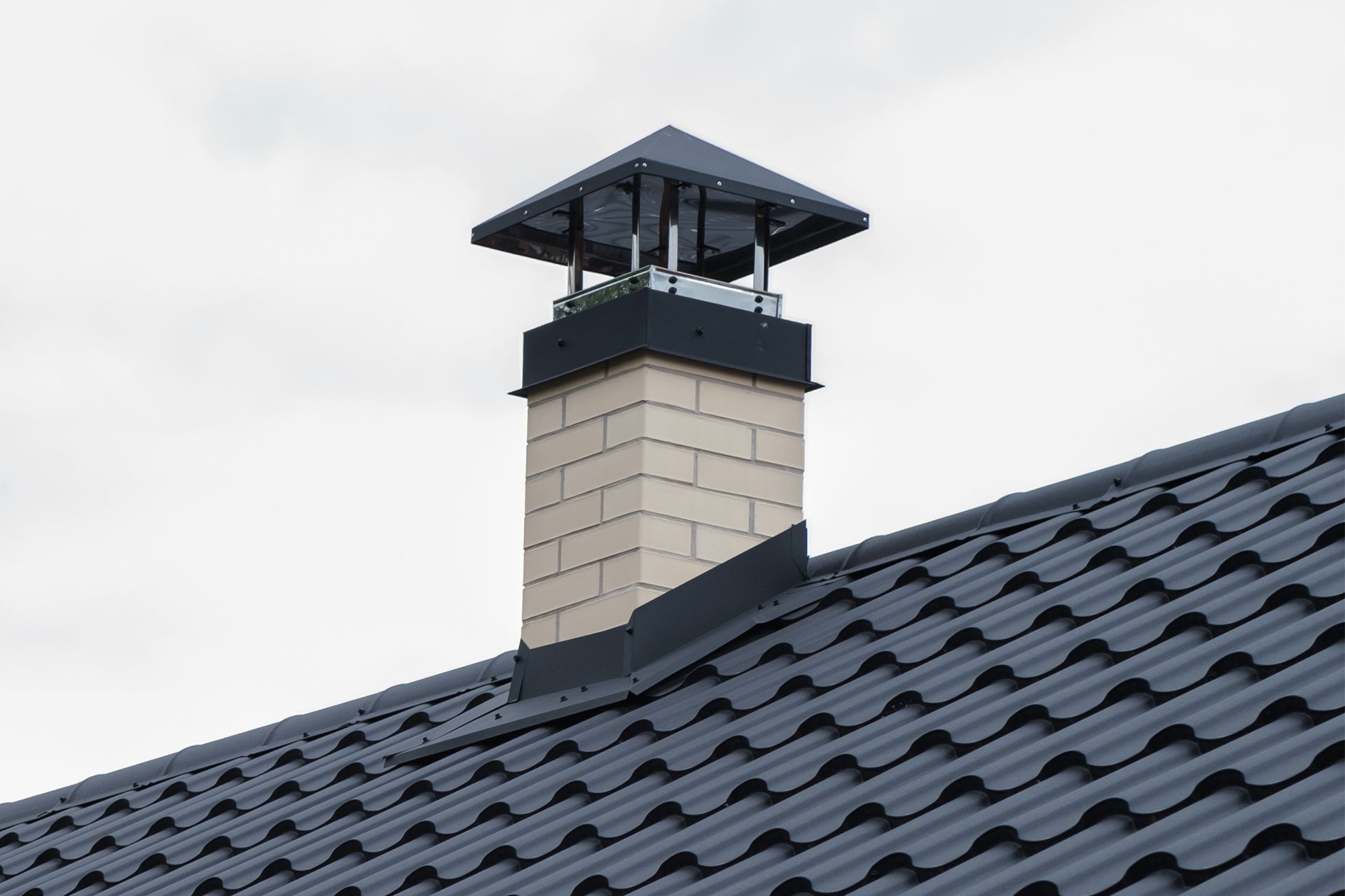 A light-colored brick chimney with a metal cap and flashing sits on a dark-colored metal shingle roof.