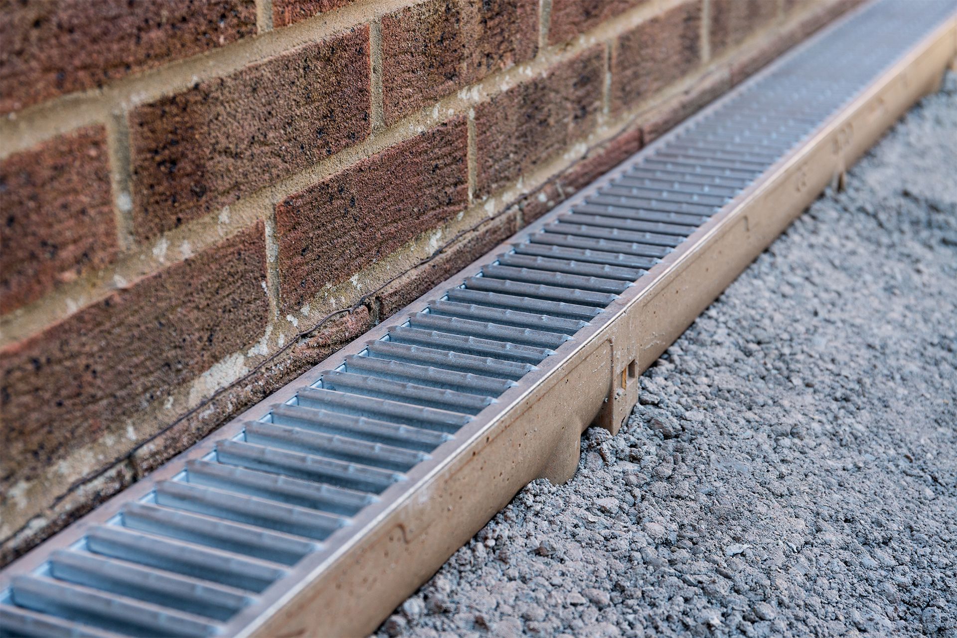 A linear drainage channel with a metal grate installed along the base of a brick wall, bordered by loose gray gravel.