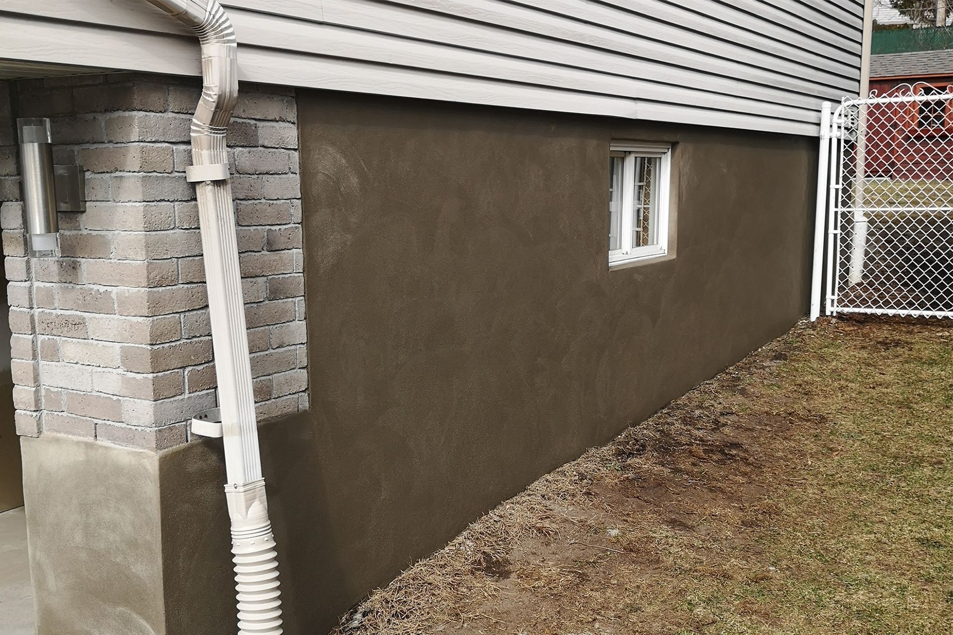 A newly parged foundation wall on a house exterior with brick accents, a downspout, and a small basement window.