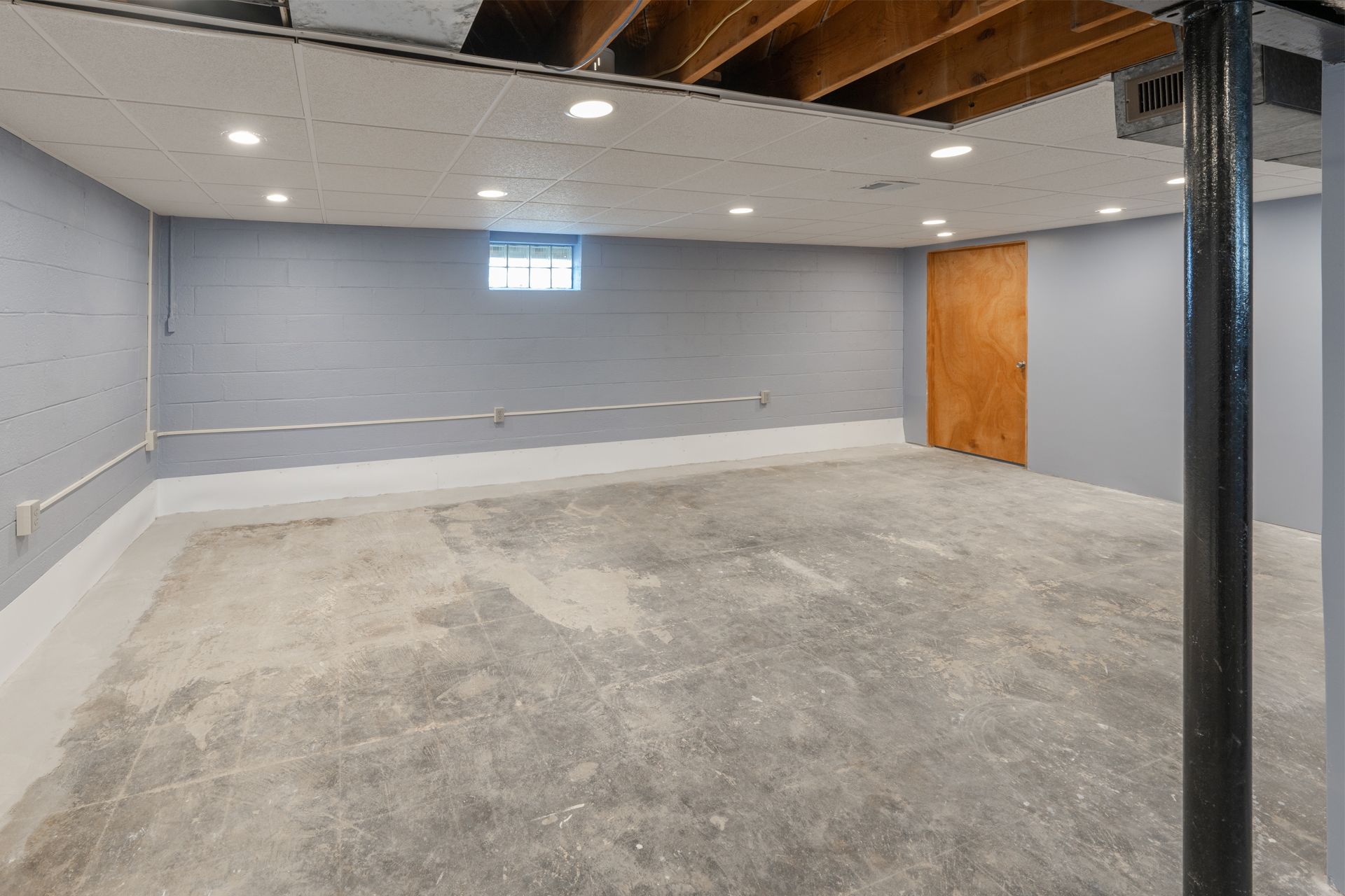 An unfinished basement with gray painted concrete block walls, a concrete floor, drop ceiling lights, and a wooden door.