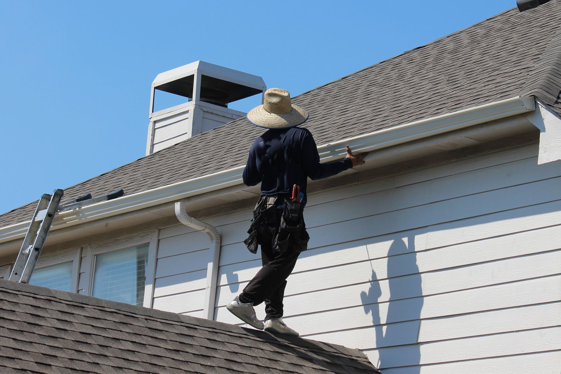A worker in a sun hat stands on a steep roof, repairing the gutters of a white-sided house under a clear blue sky.