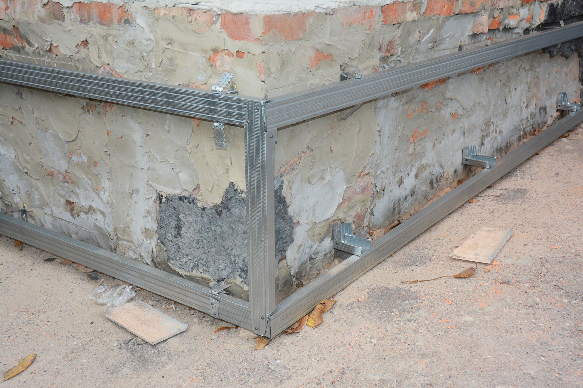 Metal framing tracks attached to the exterior brick and concrete corner of a building foundation.