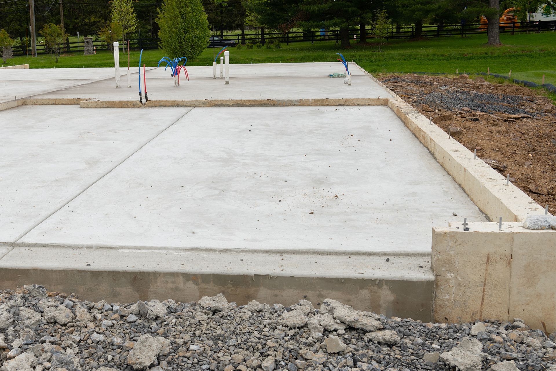 A newly poured concrete foundation slab with exposed utility pipes on a construction site with gravel and grass surroundings.