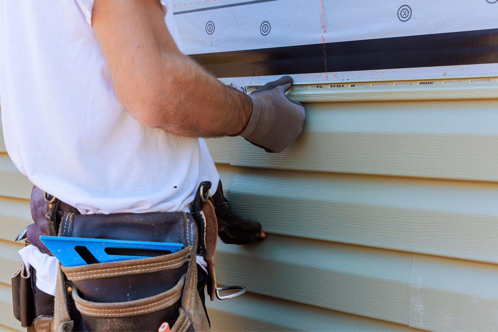 A worker in a white t-shirt and tool belt installs light green vinyl siding on a building exterior.