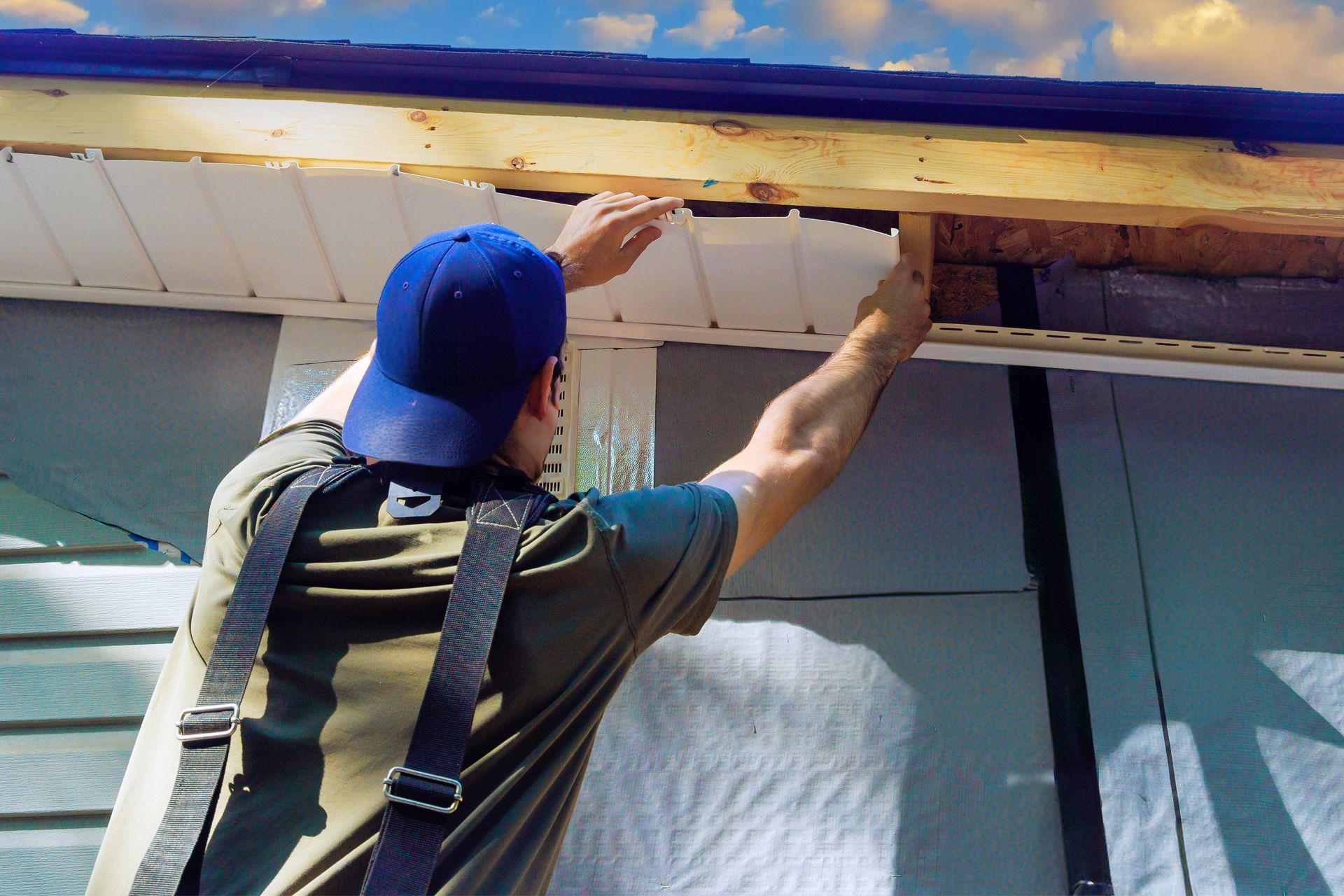 A person wearing a blue cap installs white soffit panels under the roof eaves of a house.