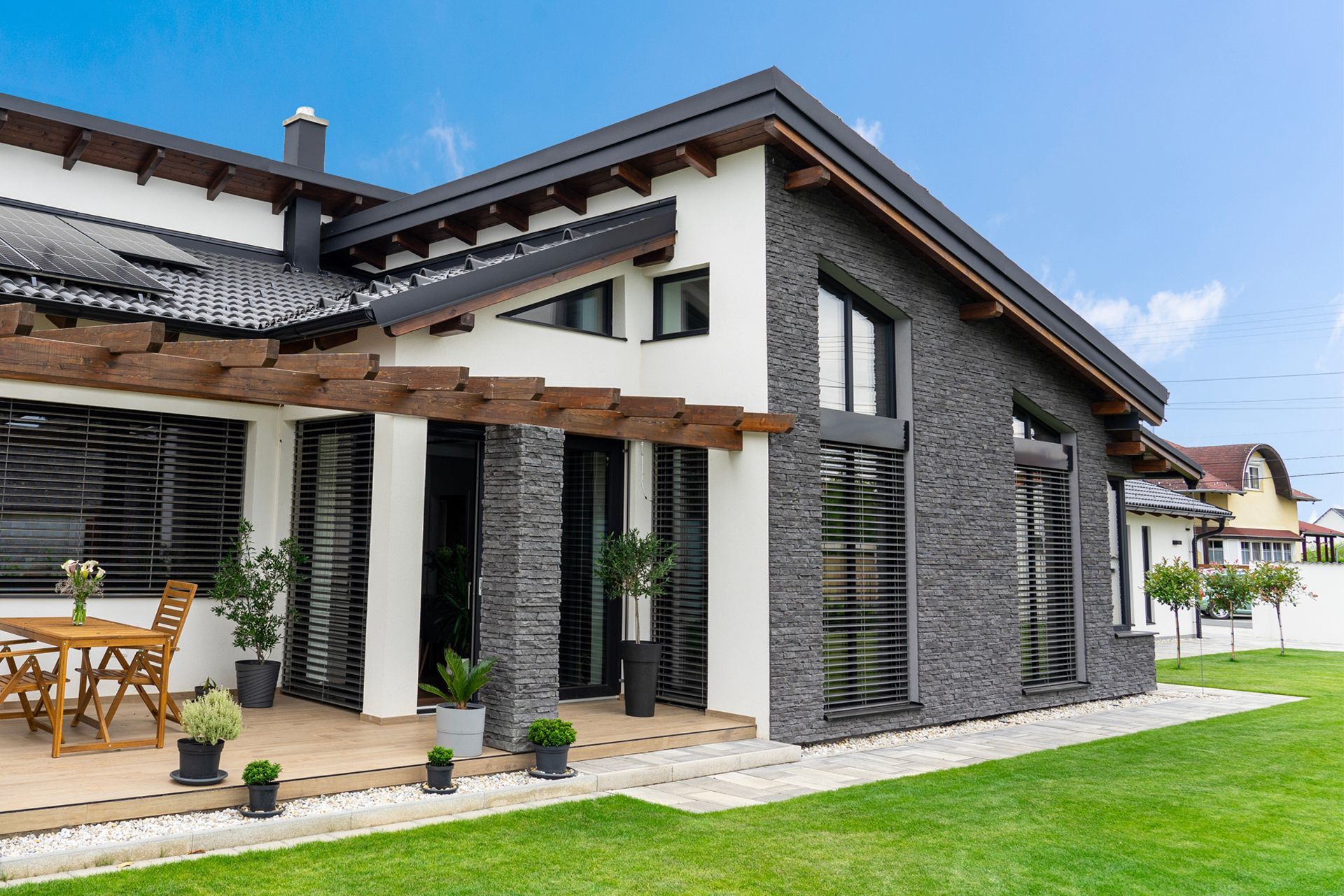 A modern house with white walls, stone accents, and a wooden pergola over a patio, surrounded by a lawn on a sunny day.