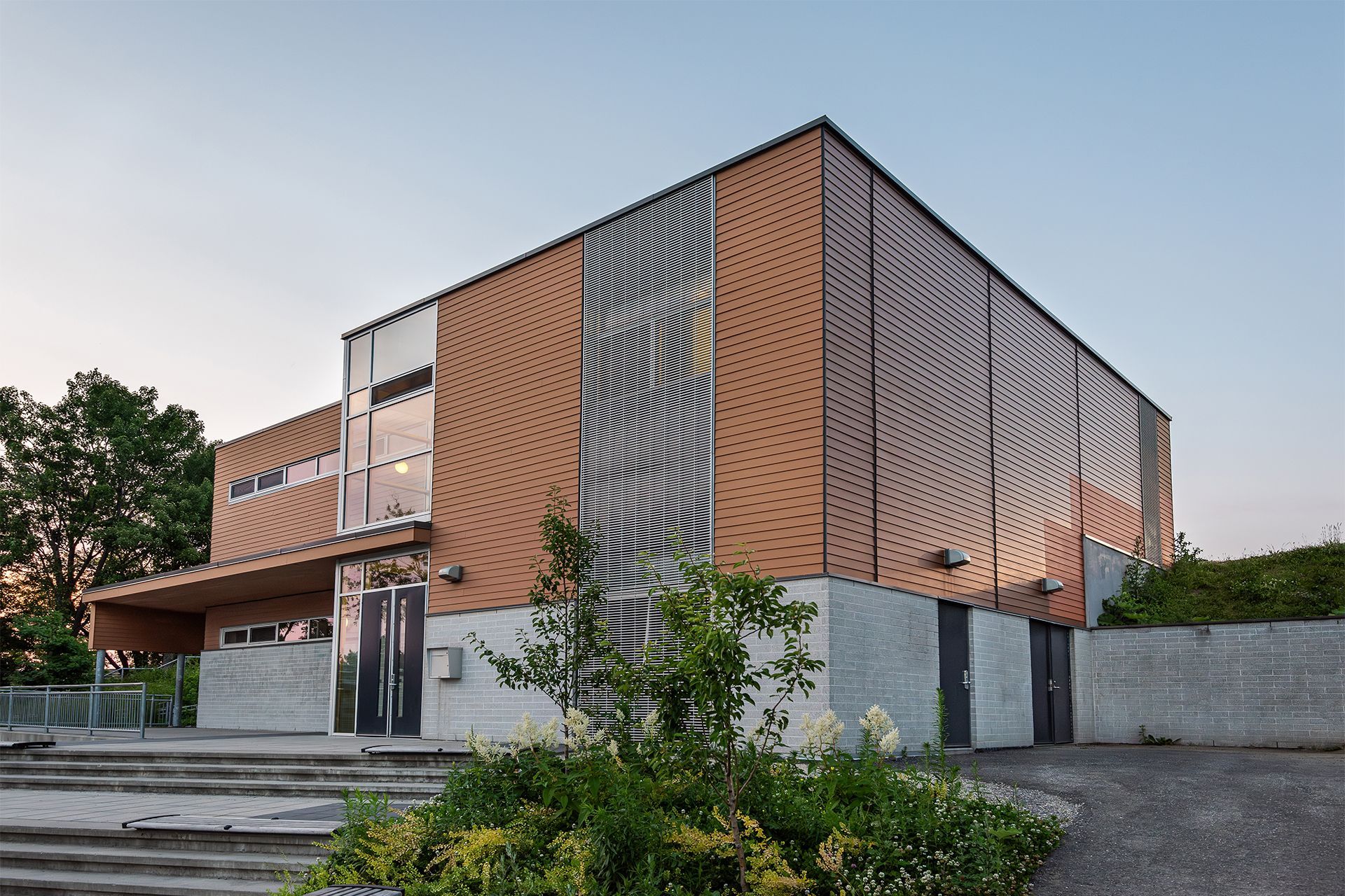 A modern two-story building with a textured, copper-colored facade and a gray concrete base, surrounded by greenery.