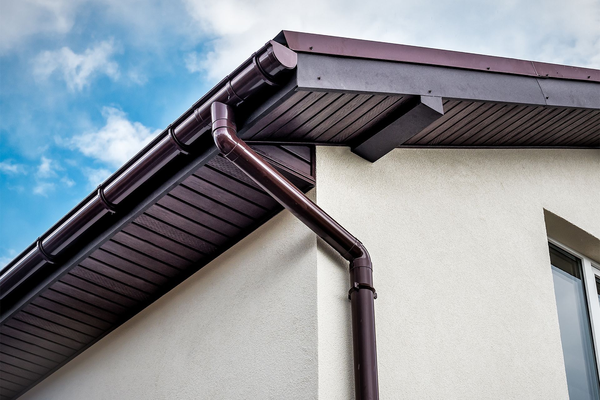 Brown gutter and downspout installed on the corner of a building with a textured off-white wall and a dark soffit.