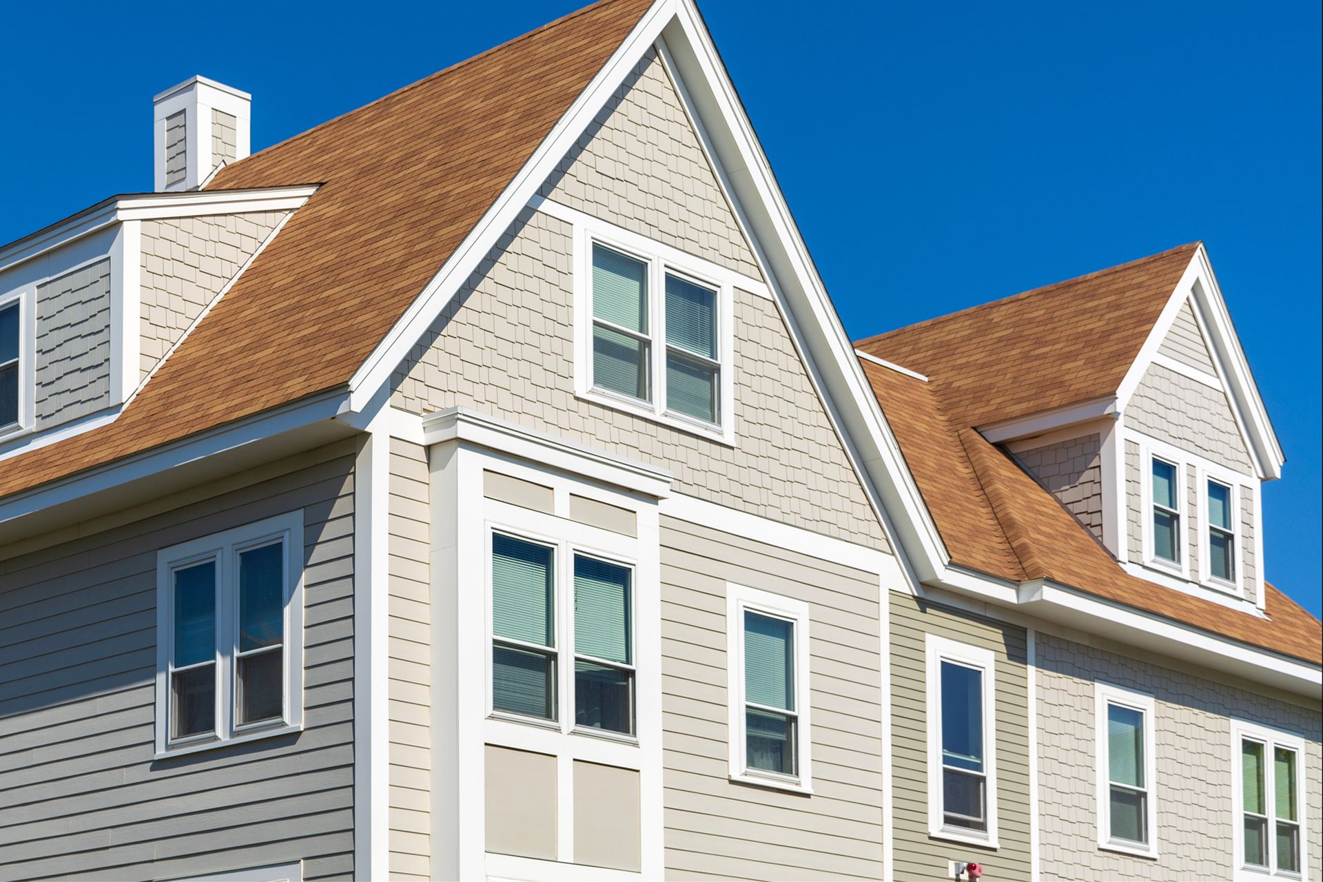 A multi-story residential building with light-colored horizontal and vertical siding, brown shingled roofs, and clear sky.