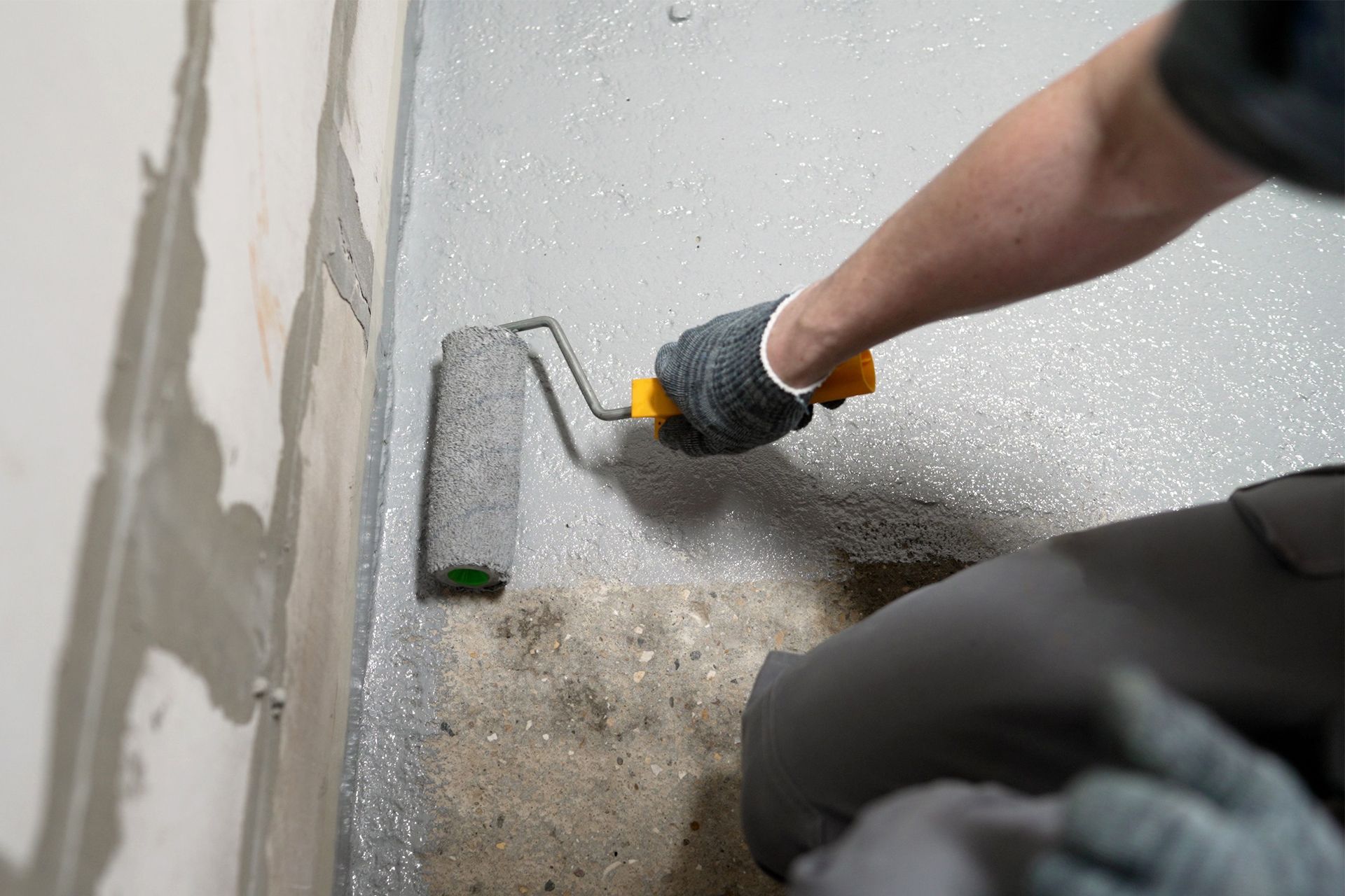 A person wearing gloves uses a paint roller to apply gray, speckled sealant to a concrete floor against a wall.