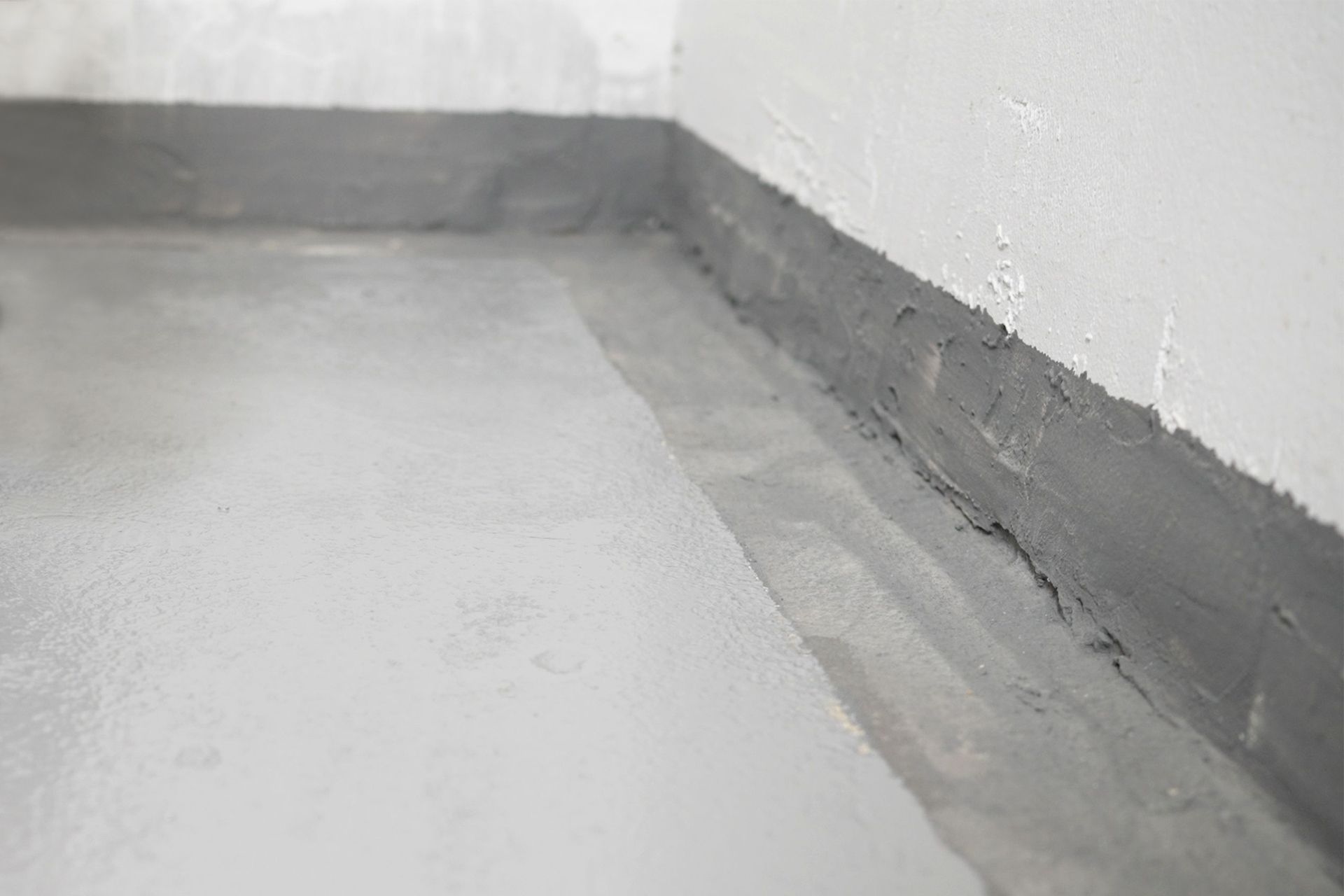 A close-up view of a floor corner where gray concrete meets a white wall with a gray waterproof sealant applied.