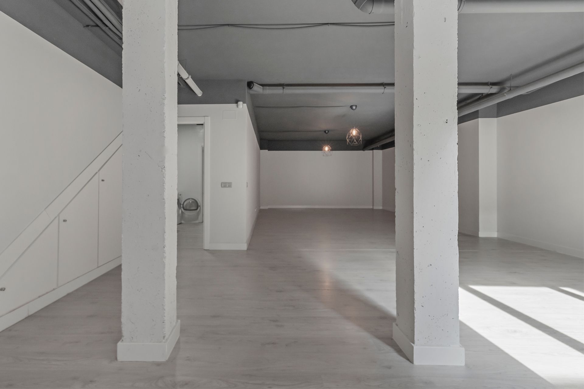 An empty, commercial basement with two white support pillars, white walls, and a grey ceiling with exposed pipes.