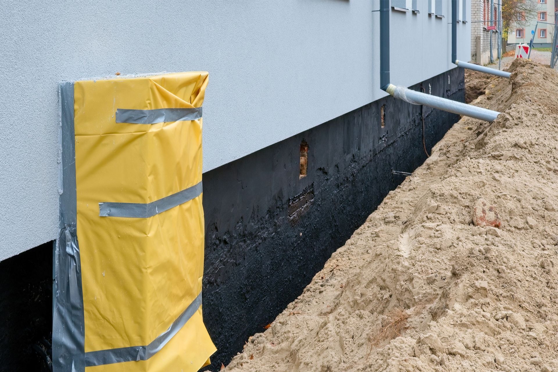 A trench along a building foundation with black waterproof coating and a section covered with yellow protective sheeting.
