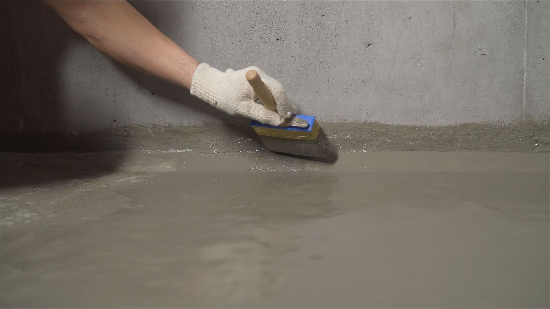A hand in a work glove uses a brush to smooth wet concrete along the corner of a gray wall.