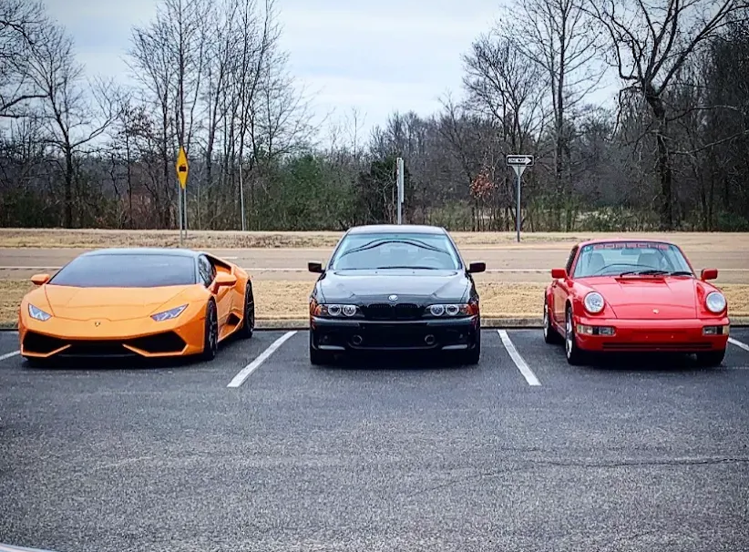 Three sports cars parked side by side in a lot: orange Lamborghini, black sedan, and red Porsche.