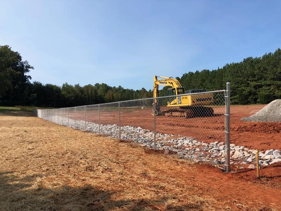 A yellow excavator is behind a chain link fence in a dirt field.